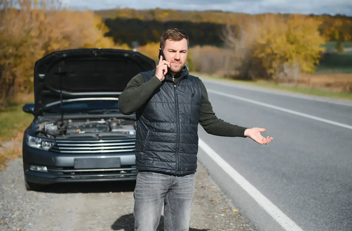 Muestra a un conductor frustrado parado junto a un auto gris oscuro con el capó abierto al costado de una carretera rural de dos carriles, haciendo una llamada telefónica.