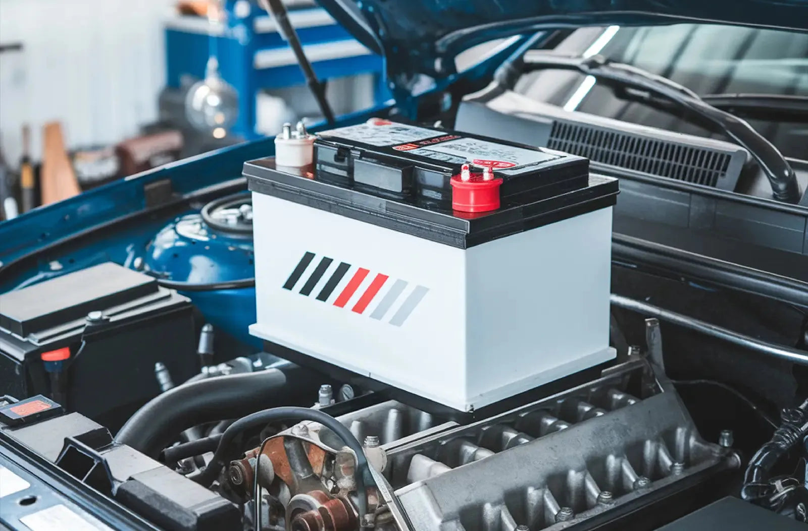 Muestra una batería de coche blanca, nueva y limpia, con protectores de terminales rojos y negros instalada dentro del compartimento del motor abierto.