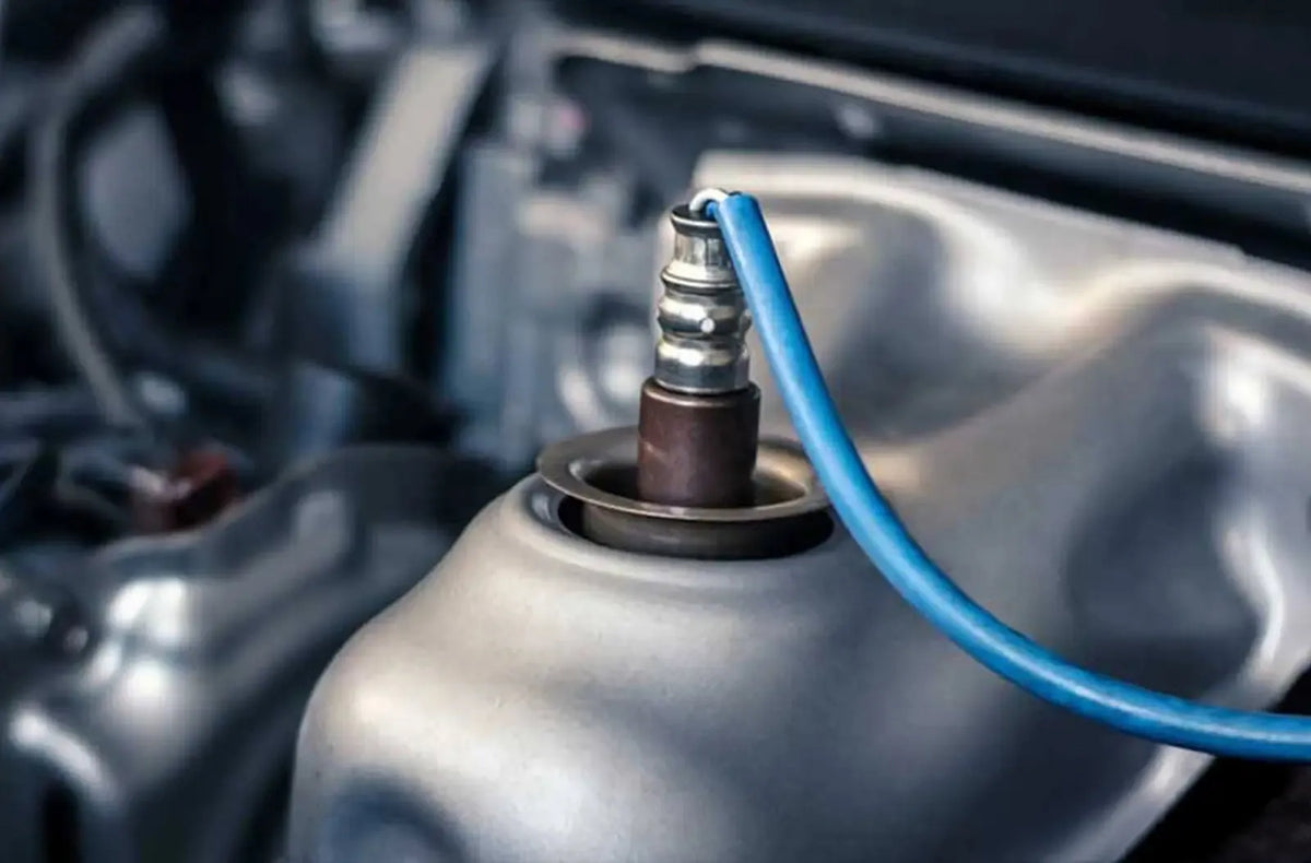 Primer plano de un sensor de oxígeno metálico y corroído instalado directamente en el tubo de escape debajo de un vehículo.