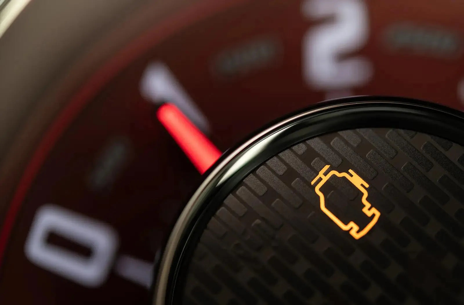 Primer plano del tablero de un vehículo, con el testigo amarillo de check engine iluminado sobre un elemento circular negro texturizado, y la aguja roja del tacómetro junto a los números desenfocados al fondo.